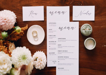 Jasne menu weselne z różowymi akcentami na stole z ciemnego drewna. Obok leżą różowe i białe kwiaty oraz obrączki.